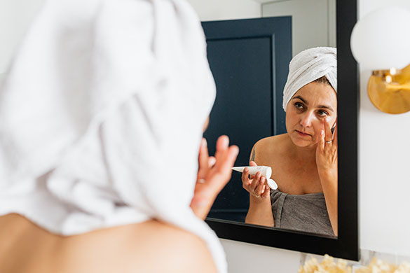 Woman with a white towel wrapped around her head applying cream to her face looking in the mirror