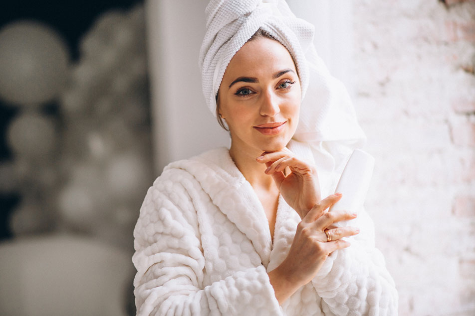 Woman in bath robe with towel wrapped around head smiling while holding lotion bottle